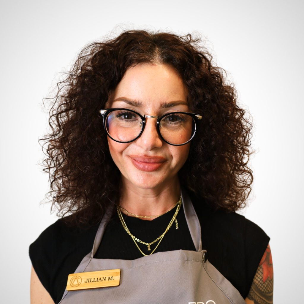 Portrait of a woman with curly brown hair and round glasses, wearing a gray apron and a name badge reading 'JILLIAN M.'