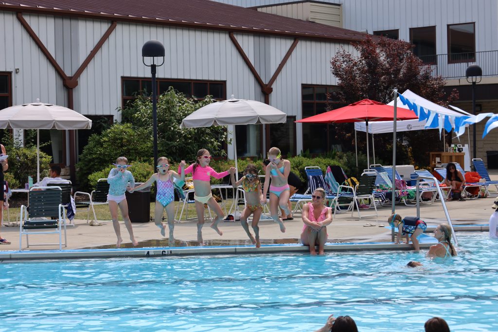 Children in swimsuits jump into a pool at a lively outdoor party with loungers and tents in the background.
