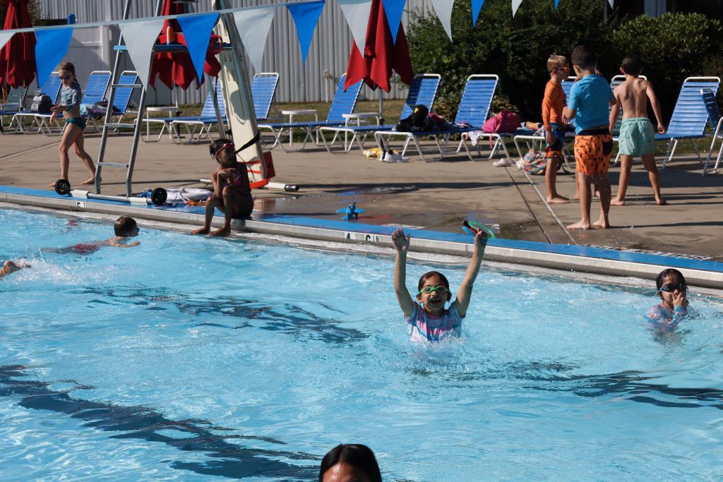 Children swimming and splashing at an outdoor pool, with blue lounge chairs and colorful bunting in the background.