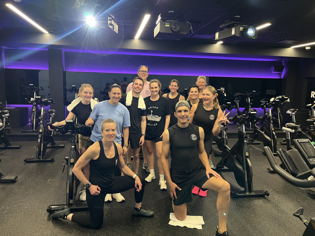 Group of people posing together in a dimly lit spin studio with stationary bikes and purple LED lighting behind them, smiling at the camera.