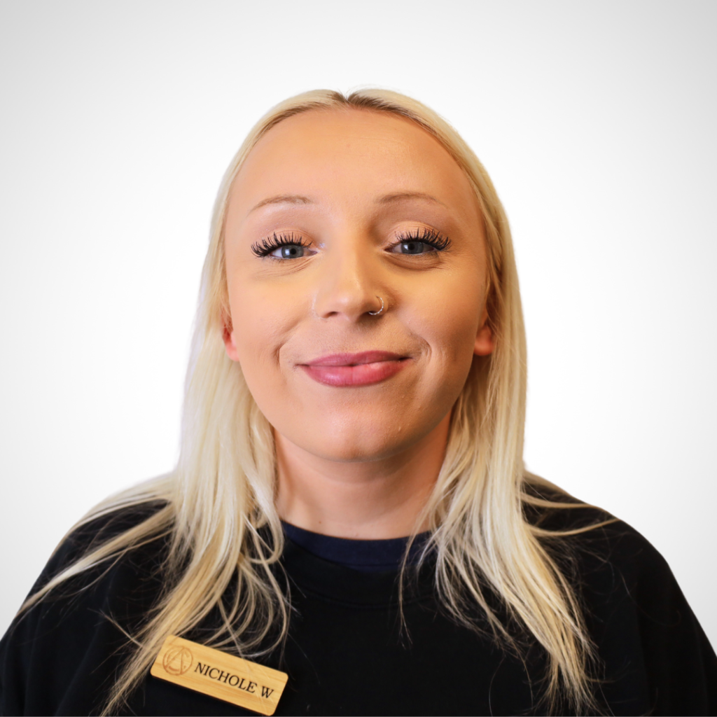 Smiling woman with blonde hair wearing a black shirt and a wooden name badge reading Nichole W, facing the camera.