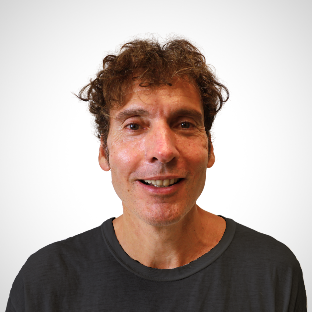 Smiling man with tousled brown hair in a dark T-shirt against a light, neutral background.
