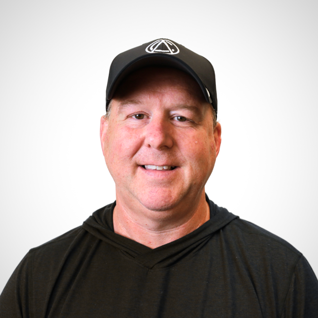 Portrait of a smiling man in a black hoodie and black cap with a white logo, against a light gray background.