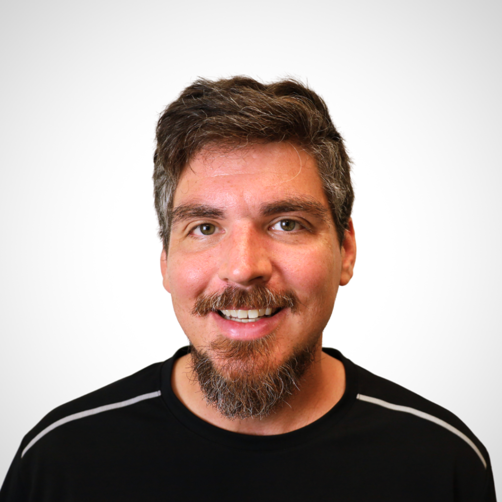 Smiling man with salt-and-pepper hair and a goatee in a head-and-shoulders portrait against a pale background, wearing a black shirt.