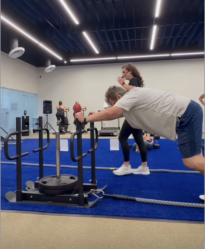 Group fitness class in a gym: a man leans forward pushing a rope pulley while a trainer or partner watches, with other participants and equipment in the background.