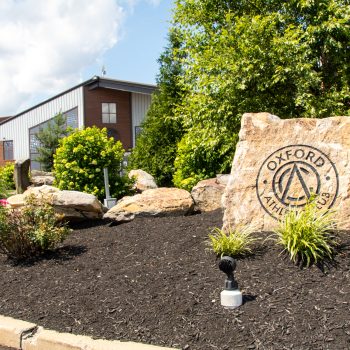 OAC_Exterior-1 Sandstone rock with Oxford Athletics emblem in a landscaped entrance area with shrubs and a modern building in the background.